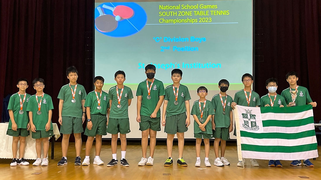 A team of students in green jerseys with silver medals pose holding a green and white striped flag, in front of a "South Zone Table Tennis Championships 2023 'C' Division Boys 2nd Position" screen.