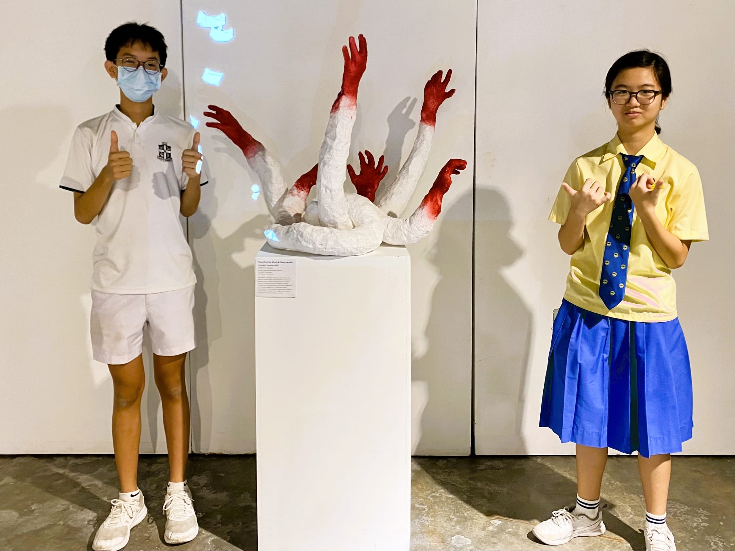 Two students in uniforms pose beside a white pedestal displaying a sculpture of red and white intertwined arm forms.