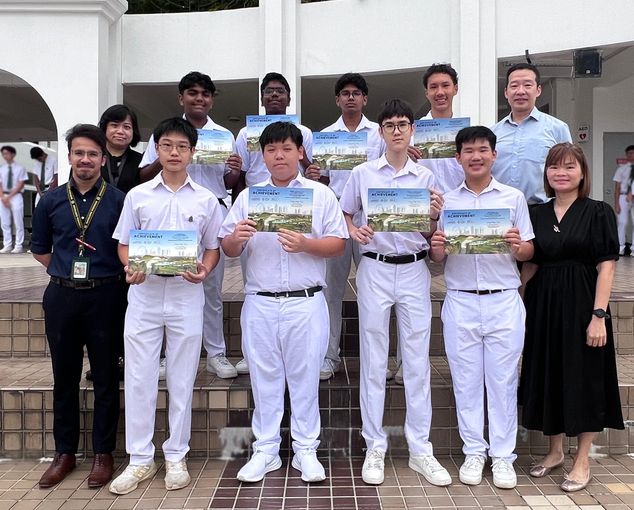 Group of students in white uniforms holding certificates of achievement, posed with several adults outdoors.
