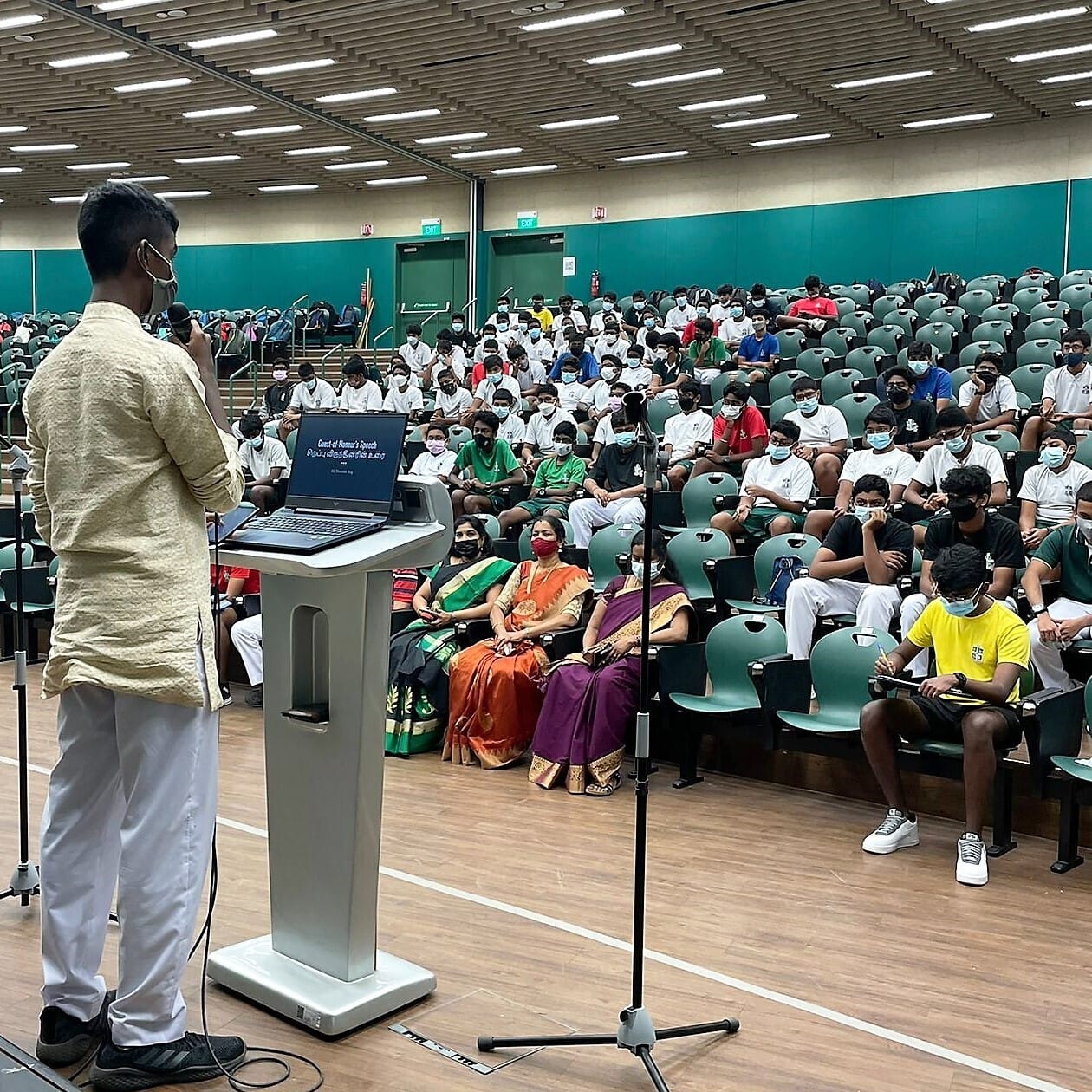 A masked speaker at a podium addresses a large seated audience of masked students in a school hall.