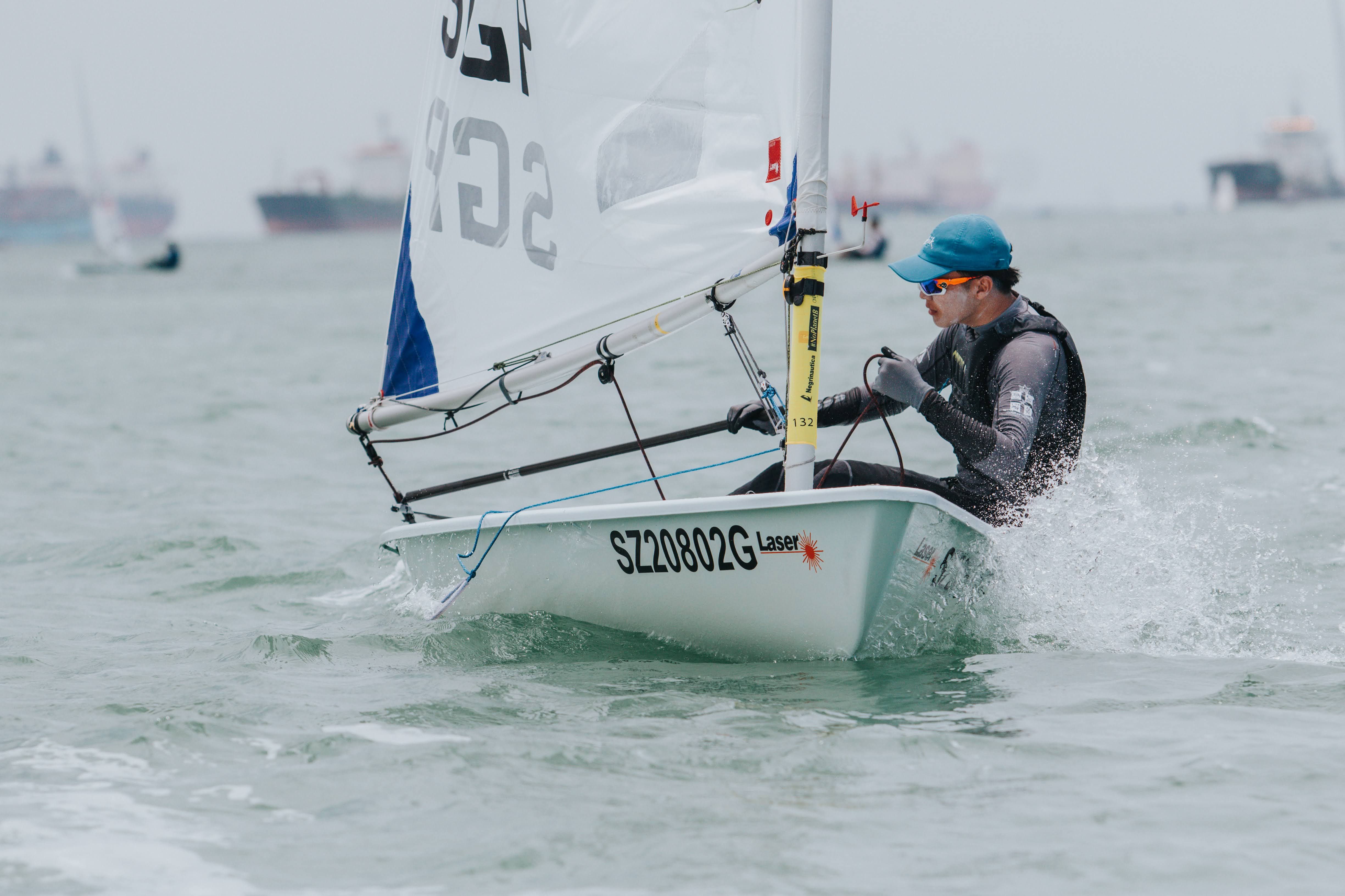 A sailor in a blue cap races a laser dinghy numbered SZ20802G, spray flying, with cargo ships in the hazy background.