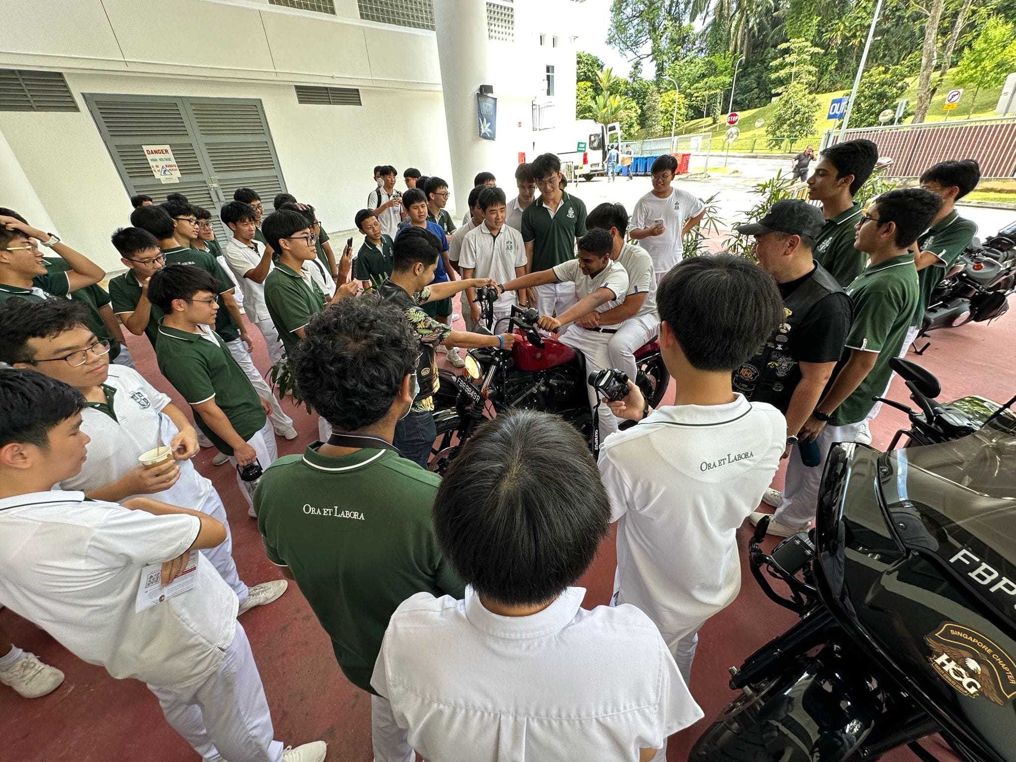 A large group of school students in green and white uniforms gather around motorcycles outdoors near a building.