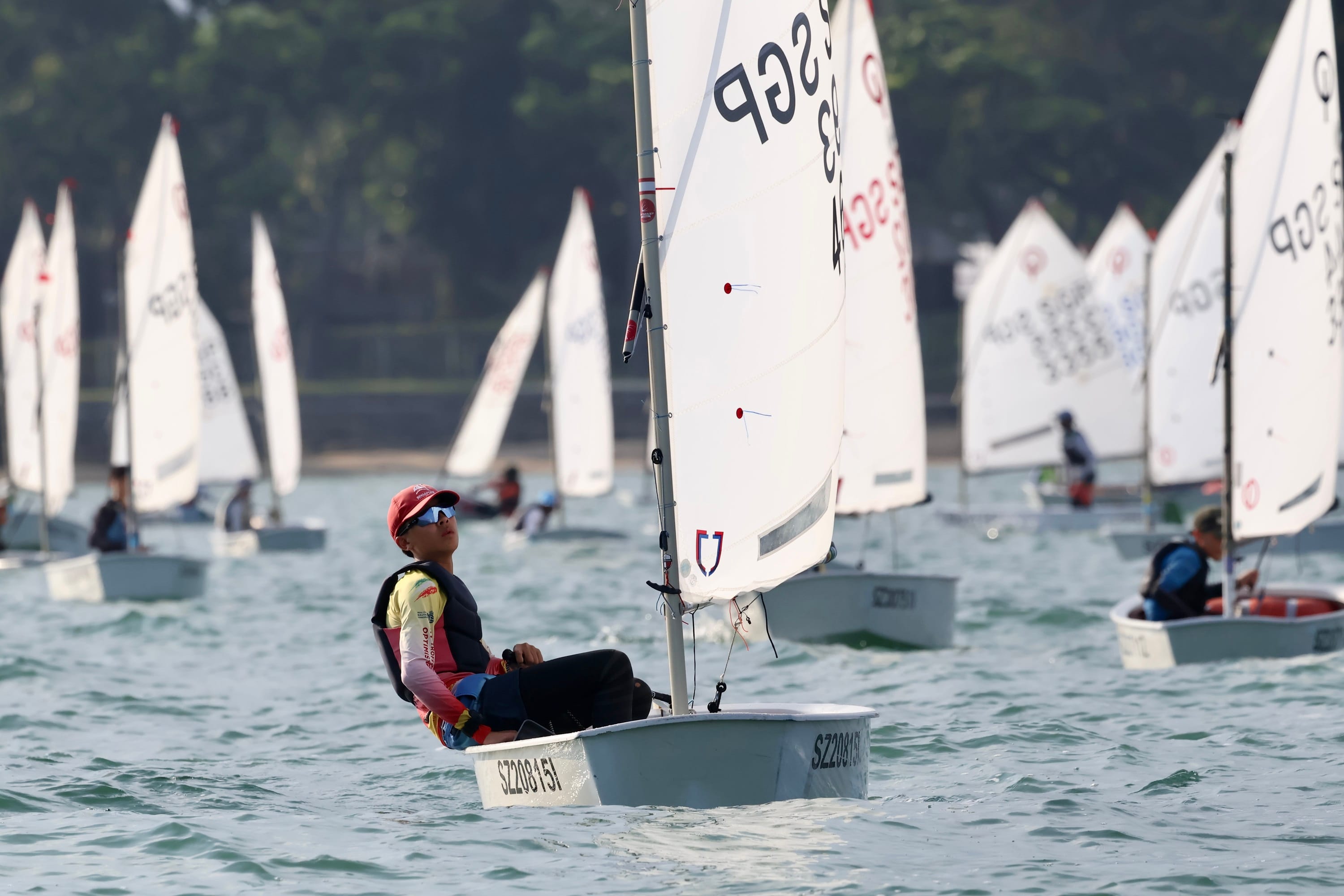 A young sailor in a red cap and sunglasses sails a small white dinghy marked SGP among many similar boats.
