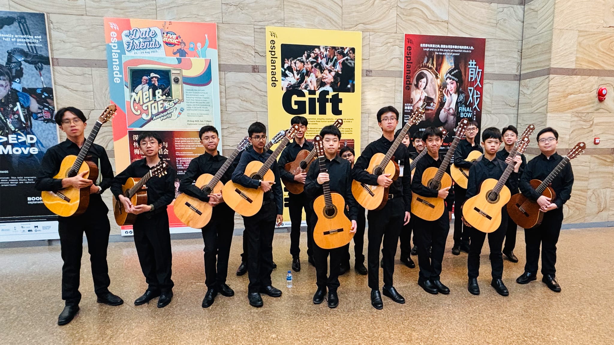 A group of young musicians in black clothing holding acoustic guitars, standing in front of Esplanade event posters.