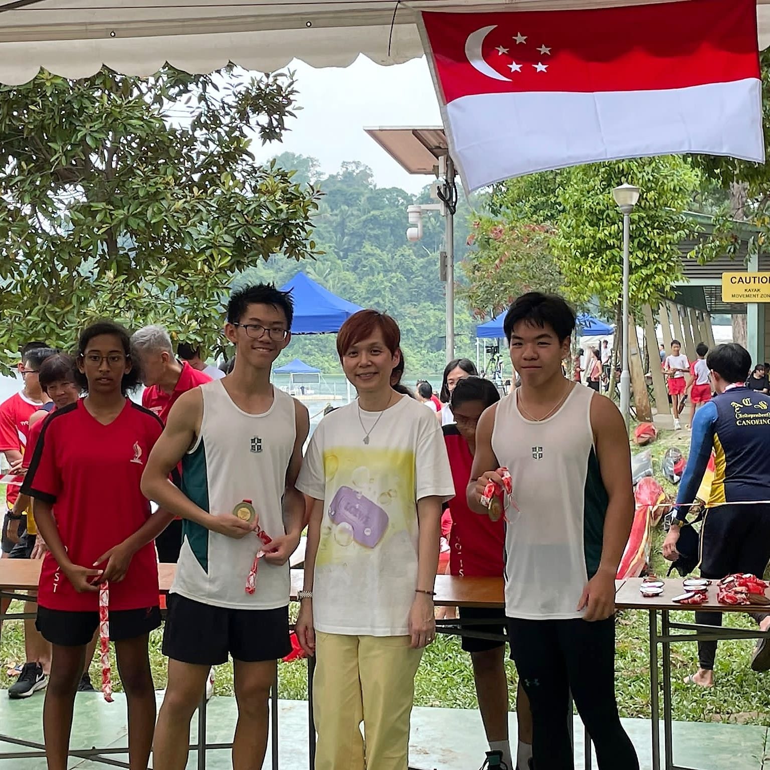 Four people pose with medals under a Singapore flag at an outdoor canoeing event near a waterway.