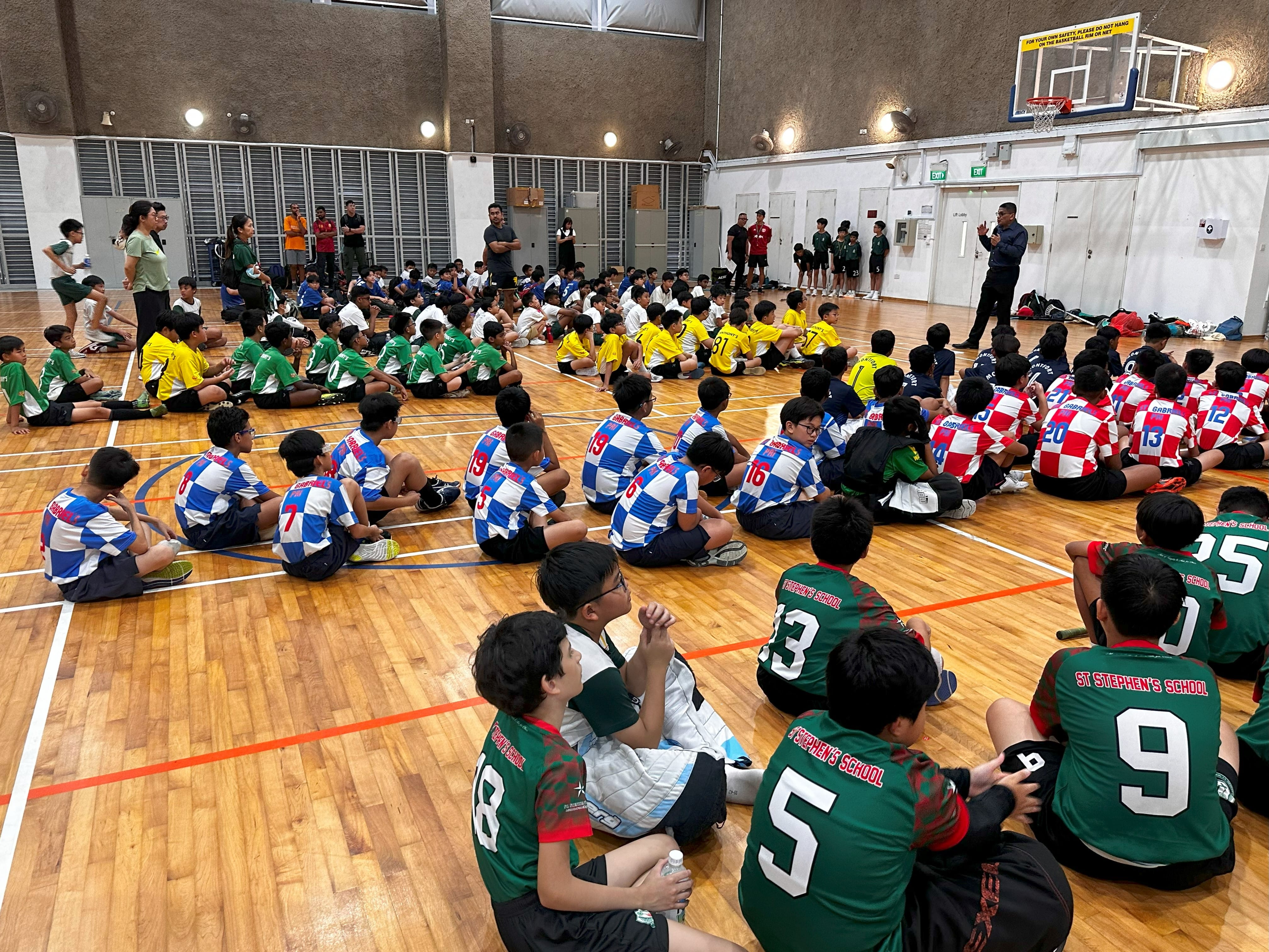 Large group of young boys in colorful sports jerseys sitting on an indoor gymnasium floor facing an adult speaker.