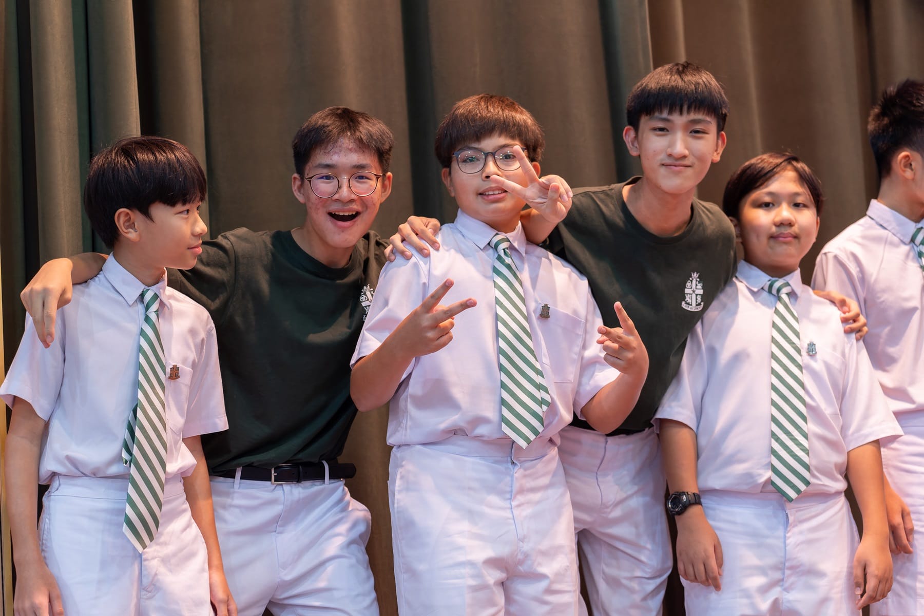 Five smiling students in white uniforms with green striped ties pose together, some making peace signs.
