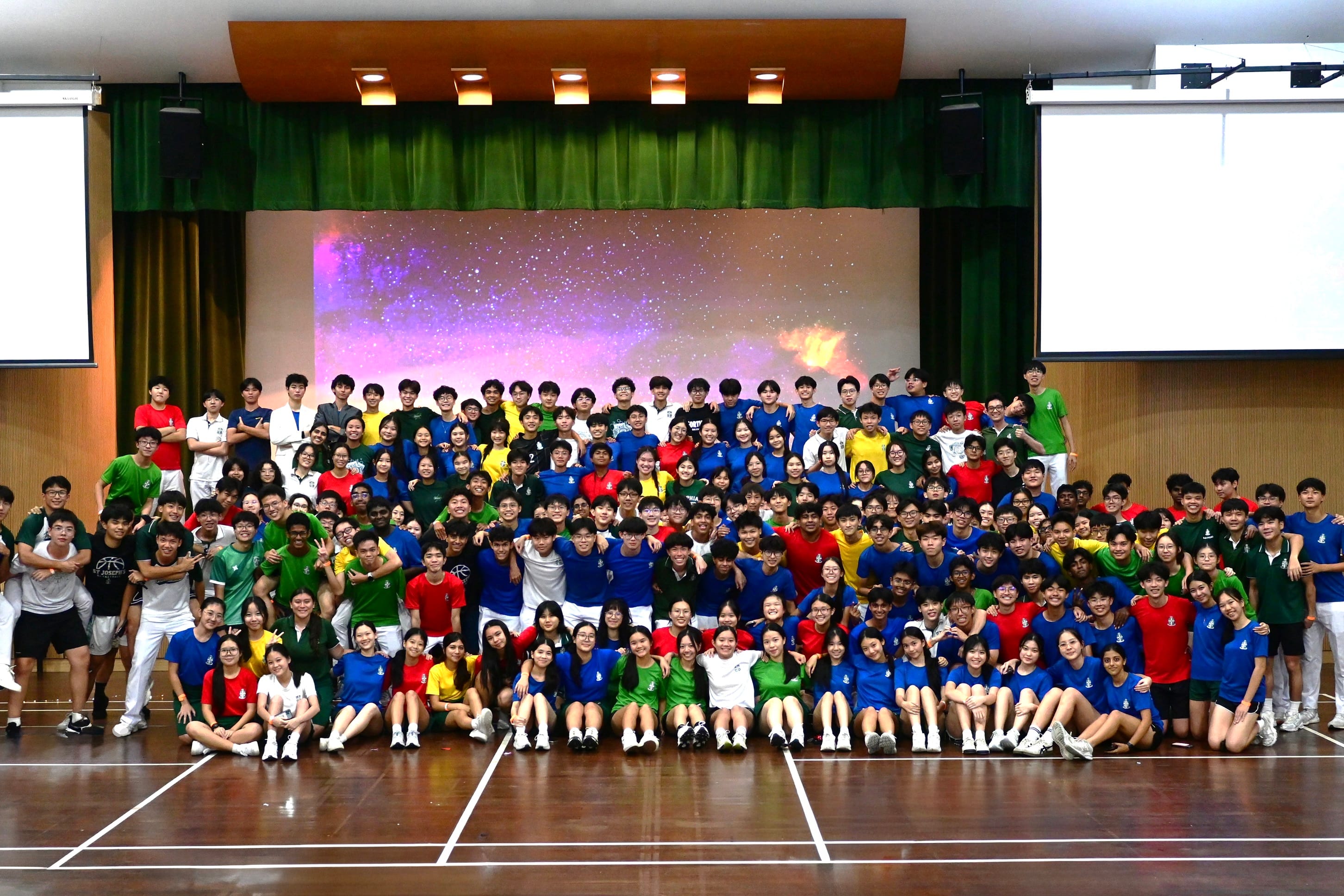 Large group of students in colorful blue, green, red, and yellow shirts posing together in a school hall.
