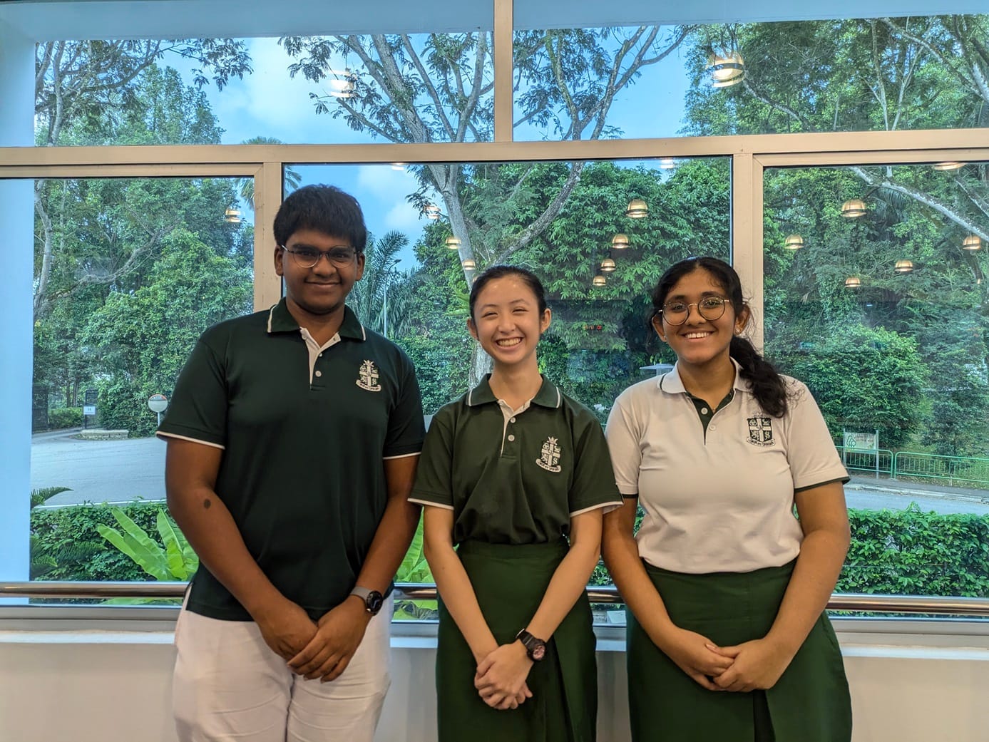 Three smiling students in green school uniforms stand together in front of large windows with trees outside.