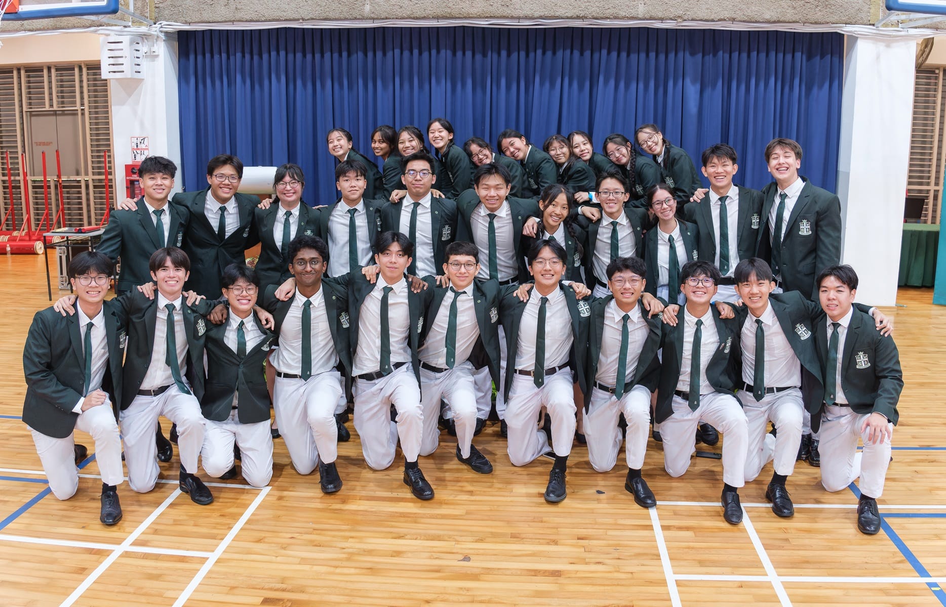 A large group of students in green blazers, white shirts, and green ties posing together in a school gymnasium.