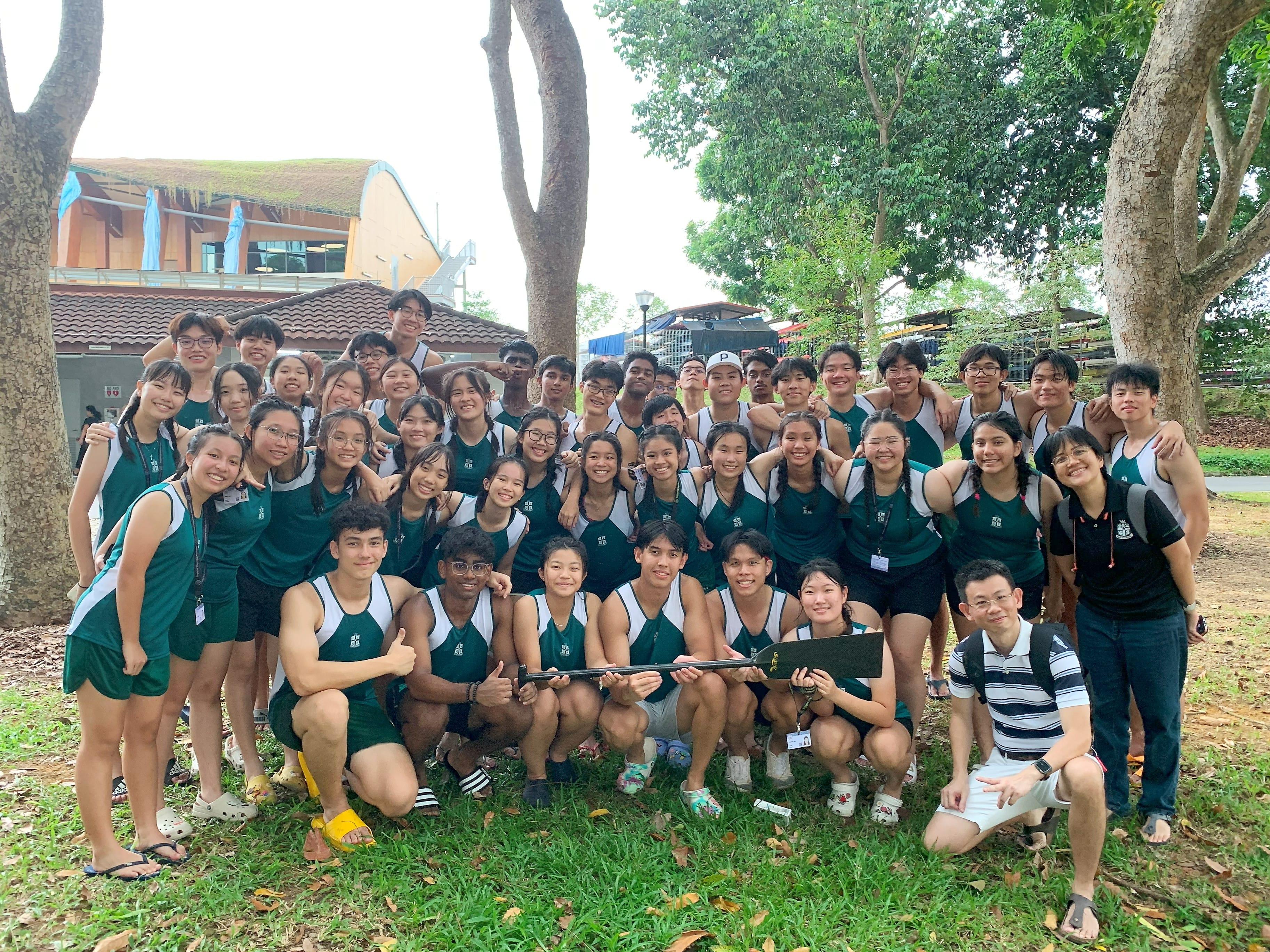 Large group of young athletes in green and white uniforms posing together on grass outdoors near trees.