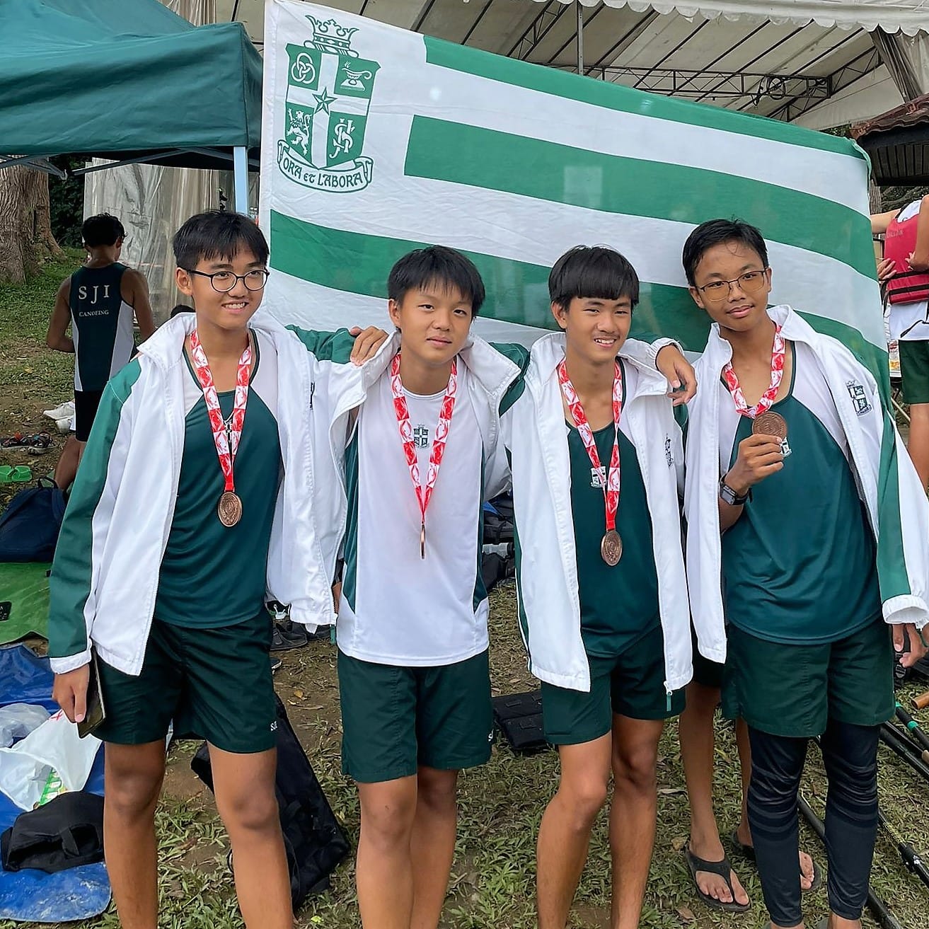 Four young male athletes in green uniforms pose with bronze medals in front of a green and white school flag.