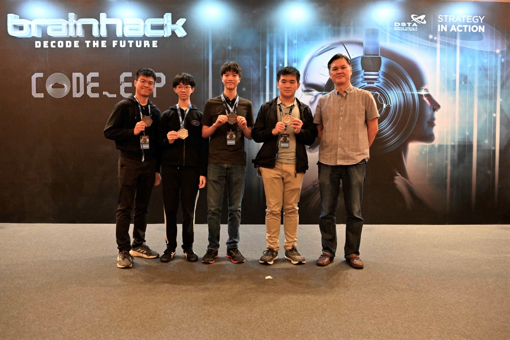 Five people pose with medals in front of a Brainhack Decode the Future event banner by DSTA.