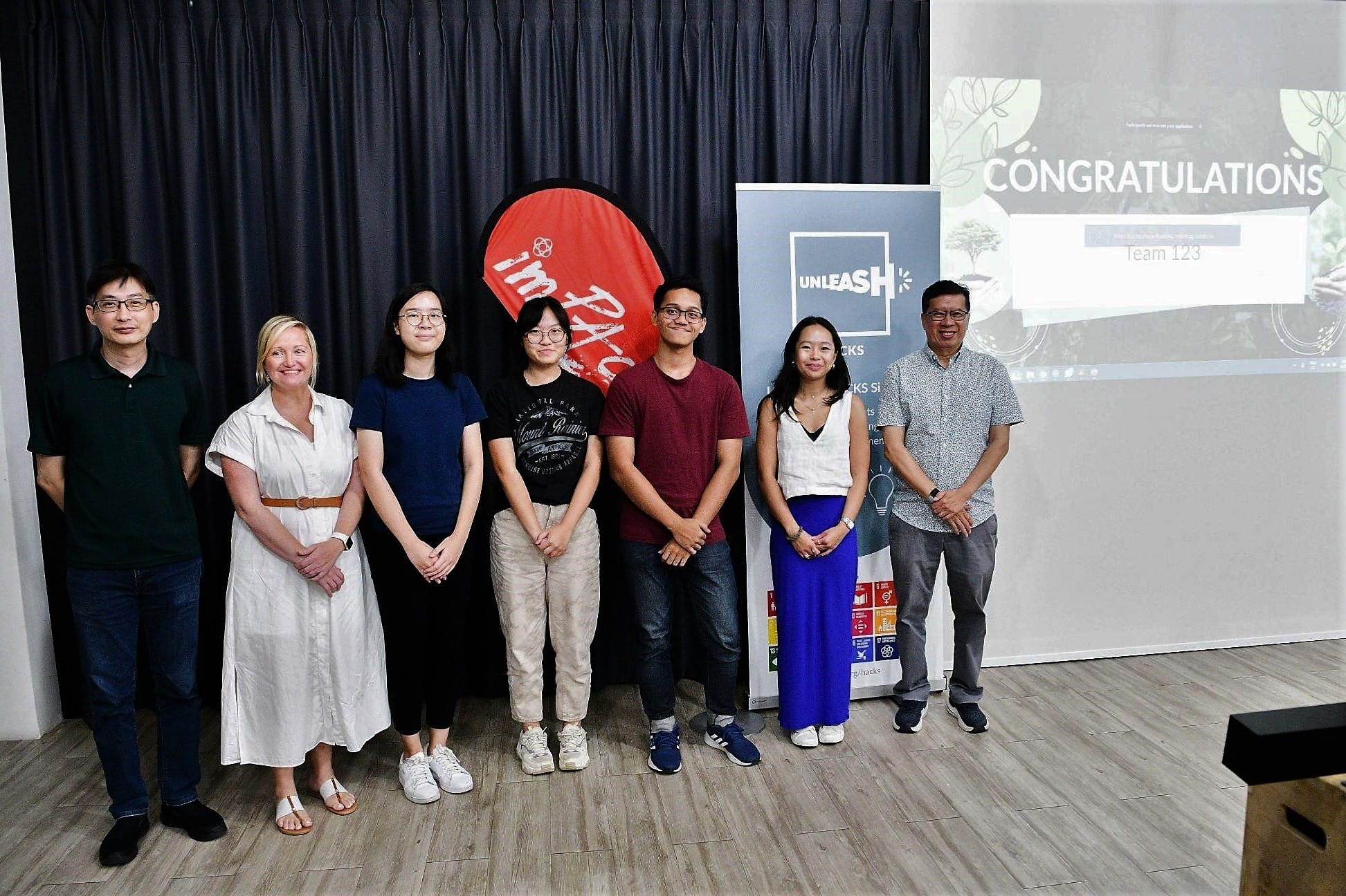 Seven people stand together near an Unleash banner and a Congratulations Team 123 projected screen.