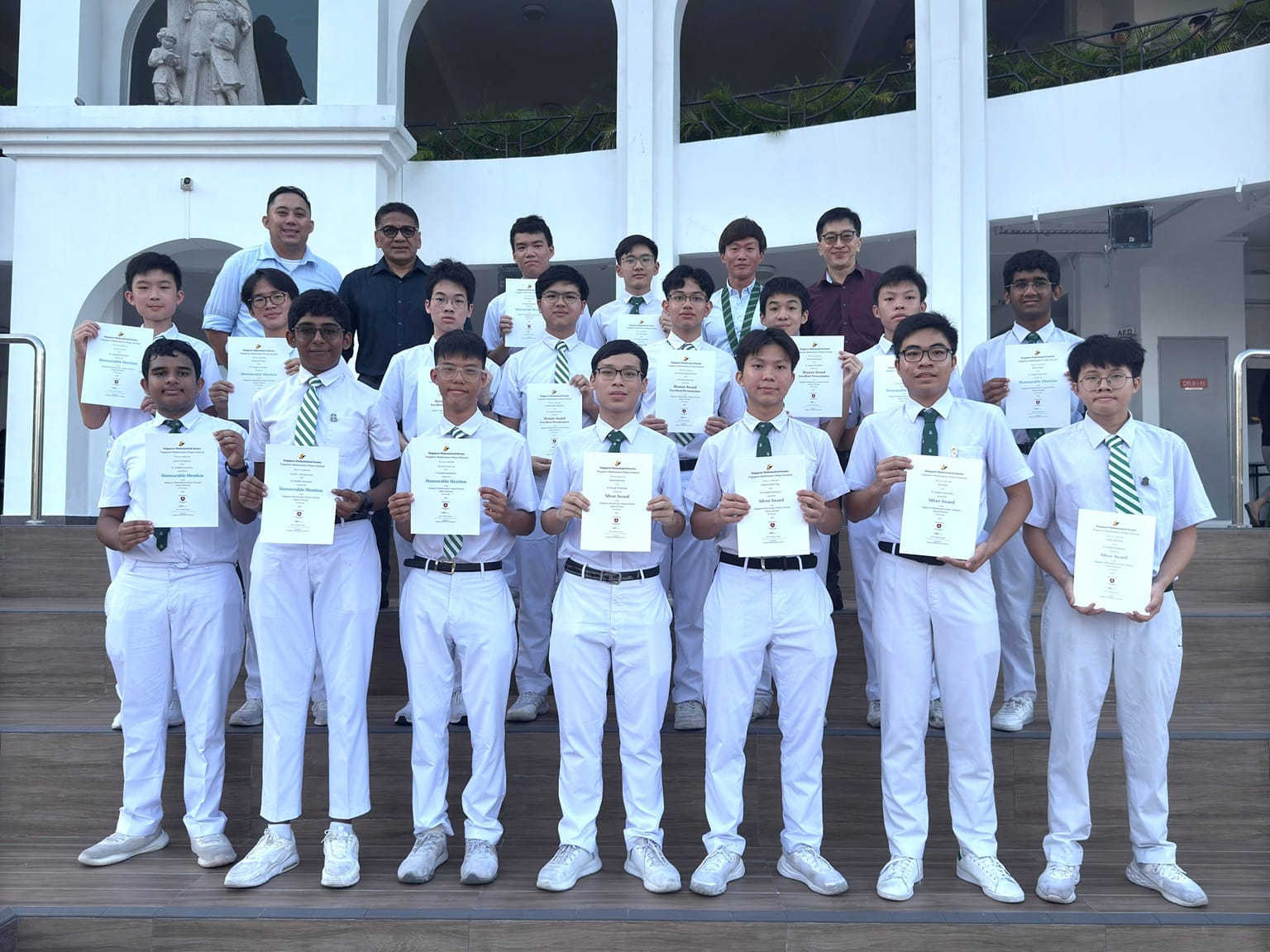A group of male students in white uniforms holding award certificates, standing on outdoor steps with teachers behind them.