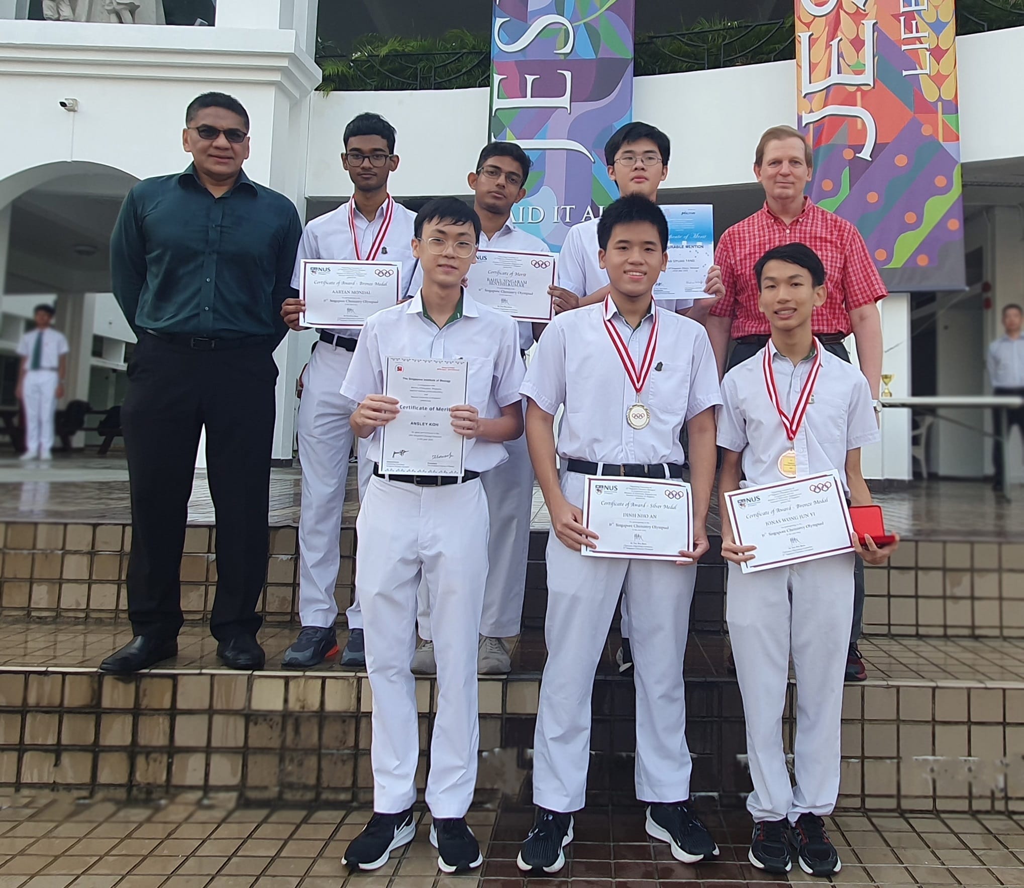 Eight people pose outdoors, students in white uniforms holding Chemistry Olympiad certificates and medals with two adults.