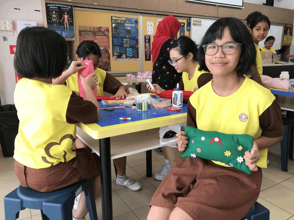 Girl with glasses holds a green embroidered pillow while classmates do crafts at a table in a classroom.
