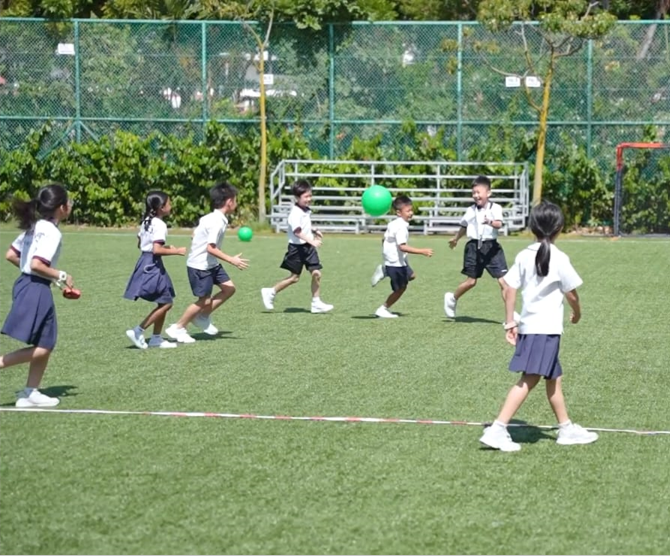 Children in school uniforms playing with a green ball on a grassy field enclosed by a chain-link fence.
