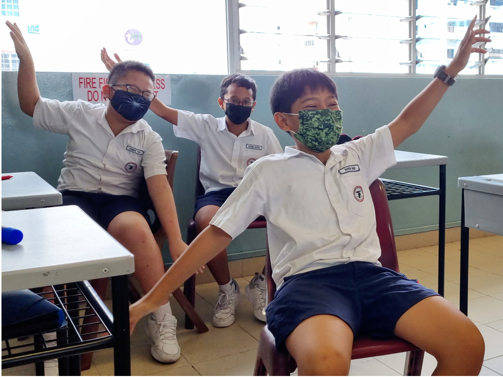 Three masked male students in white uniforms sit in a classroom with arms raised enthusiastically.