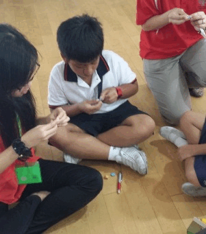 Three children sit on a wooden floor, handling small objects, with one boy in a white shirt cross-legged in the center.