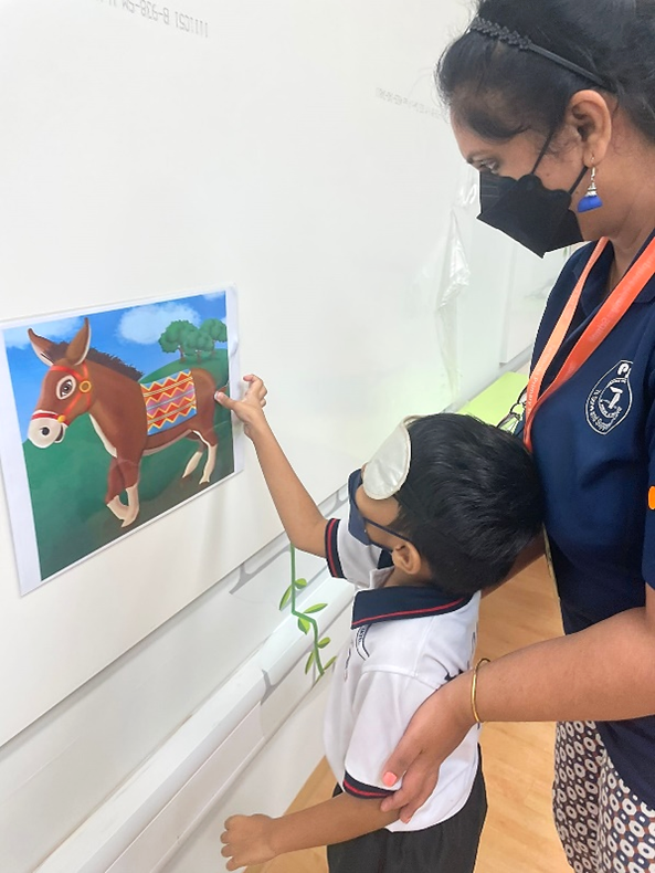 A masked teacher helps a young student point at a colorful donkey illustration on a white wall.