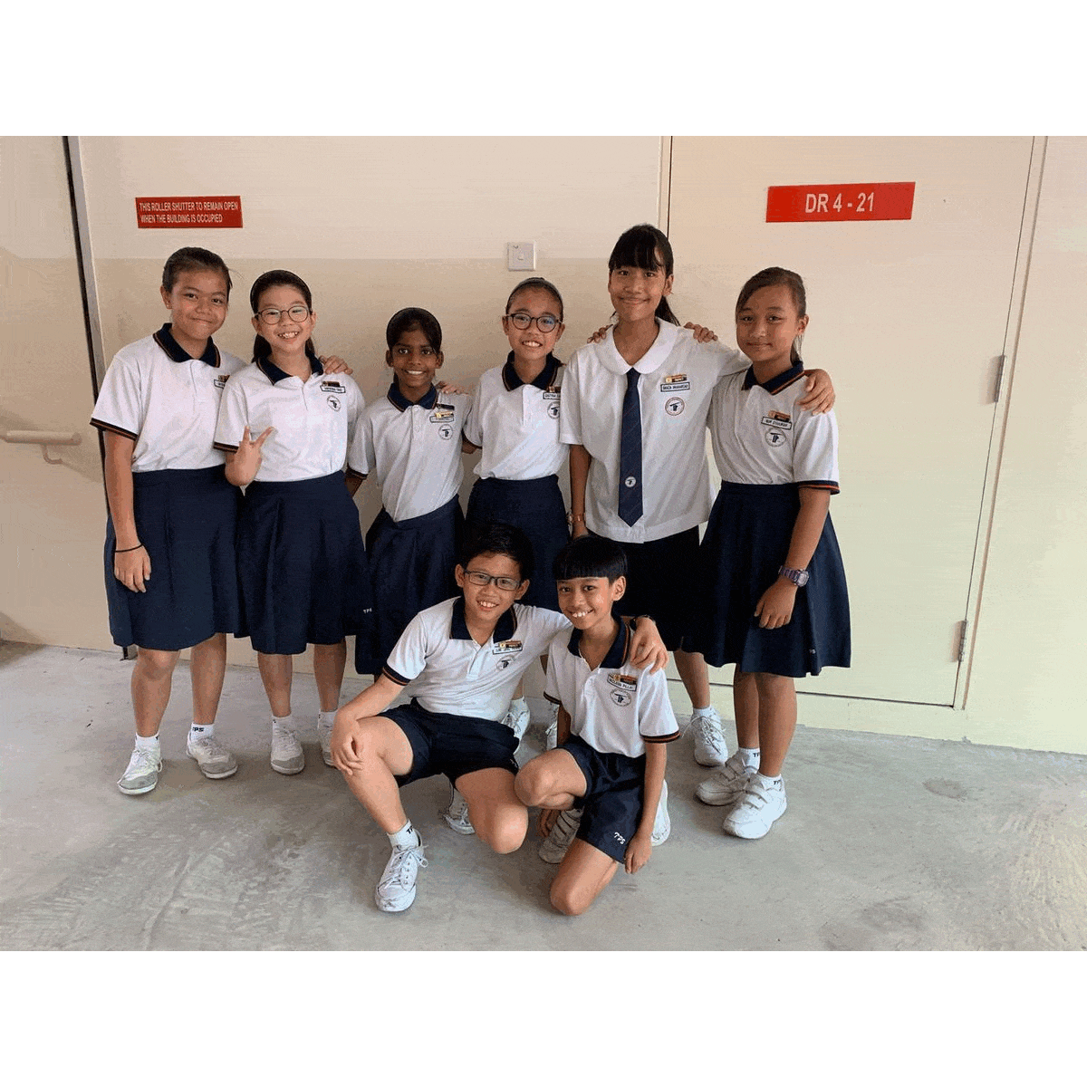 Eight students in white school uniforms pose together in a hallway by a door marked DR 4-21
