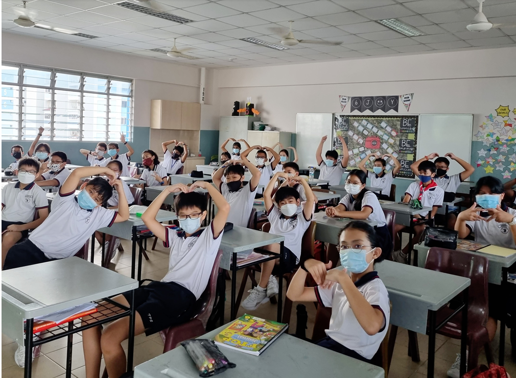 Students in a classroom wearing masks form heart shapes with their hands while seated at desks.