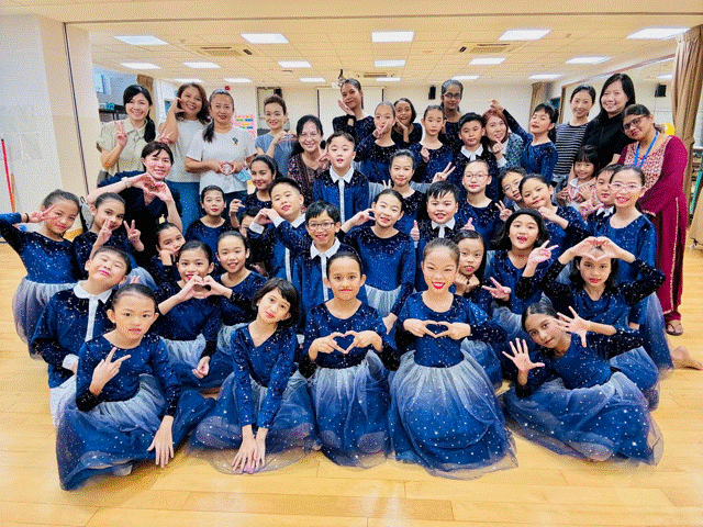 A large group of children in matching blue sparkly dance costumes pose with adults, making heart and peace signs.