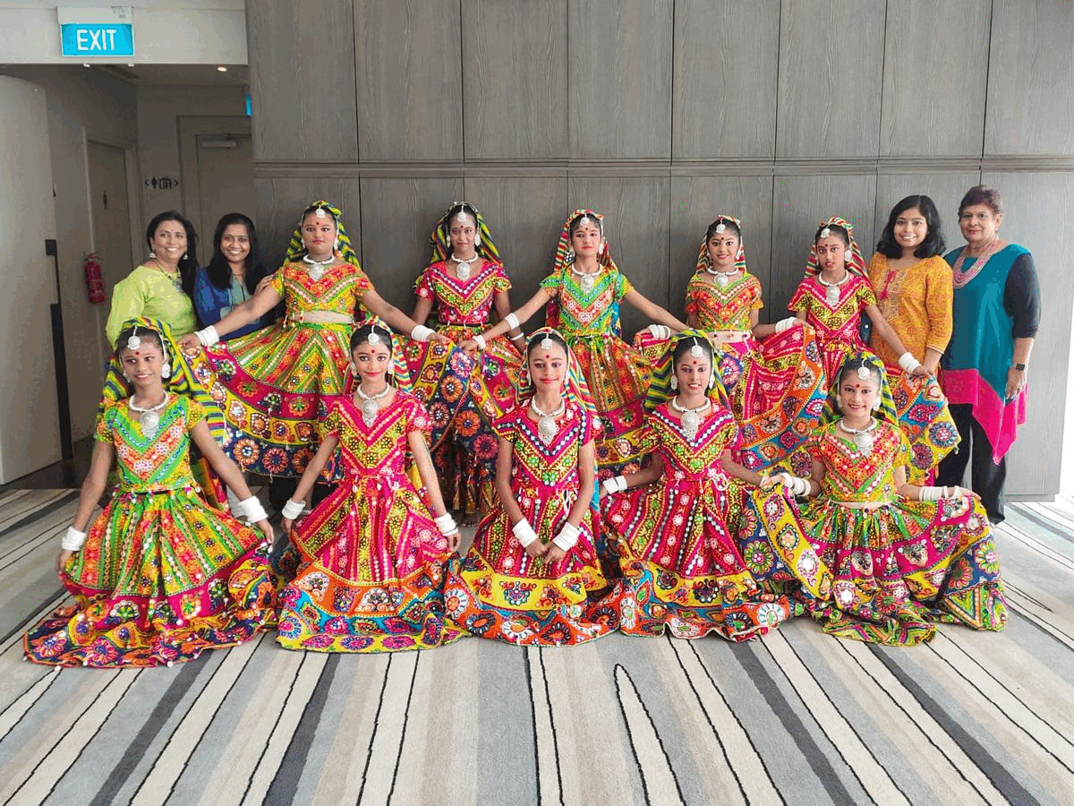 Group of girls in vibrant embroidered Indian Garba dance costumes posing indoors with two women standing behind.