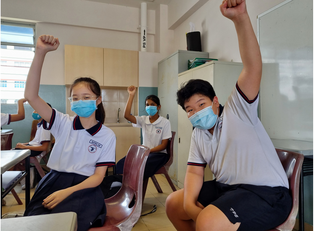Four students in school uniforms and blue face masks raise their fists in a classroom.