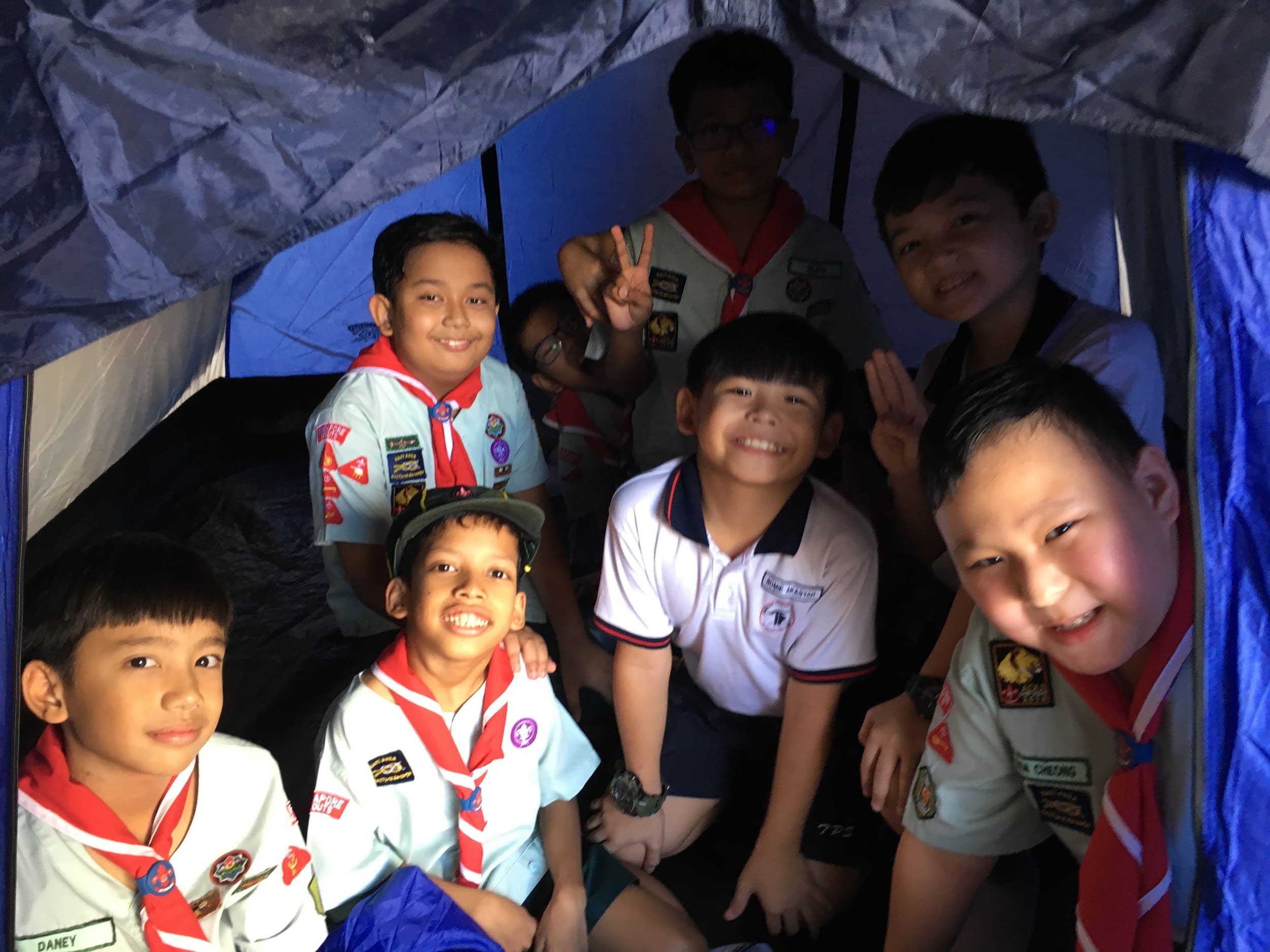 A group of young boys in scout uniforms with badges and red neckerchiefs smiling inside a blue tent.