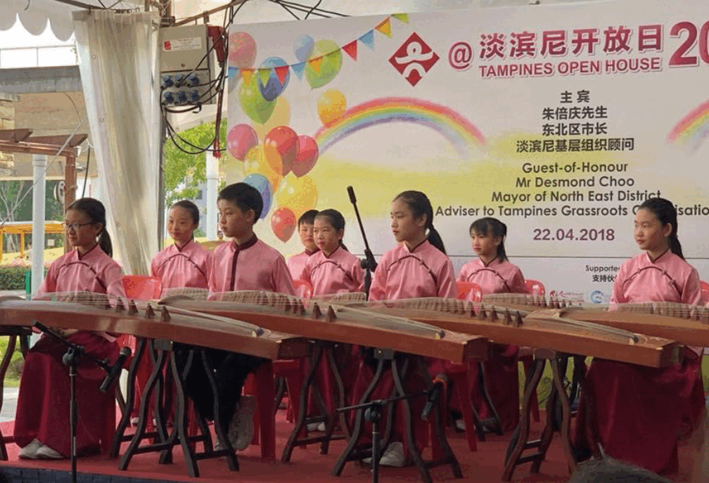 Seven children in pink outfits playing guzheng instruments on a stage at Tampines Open House 2018.