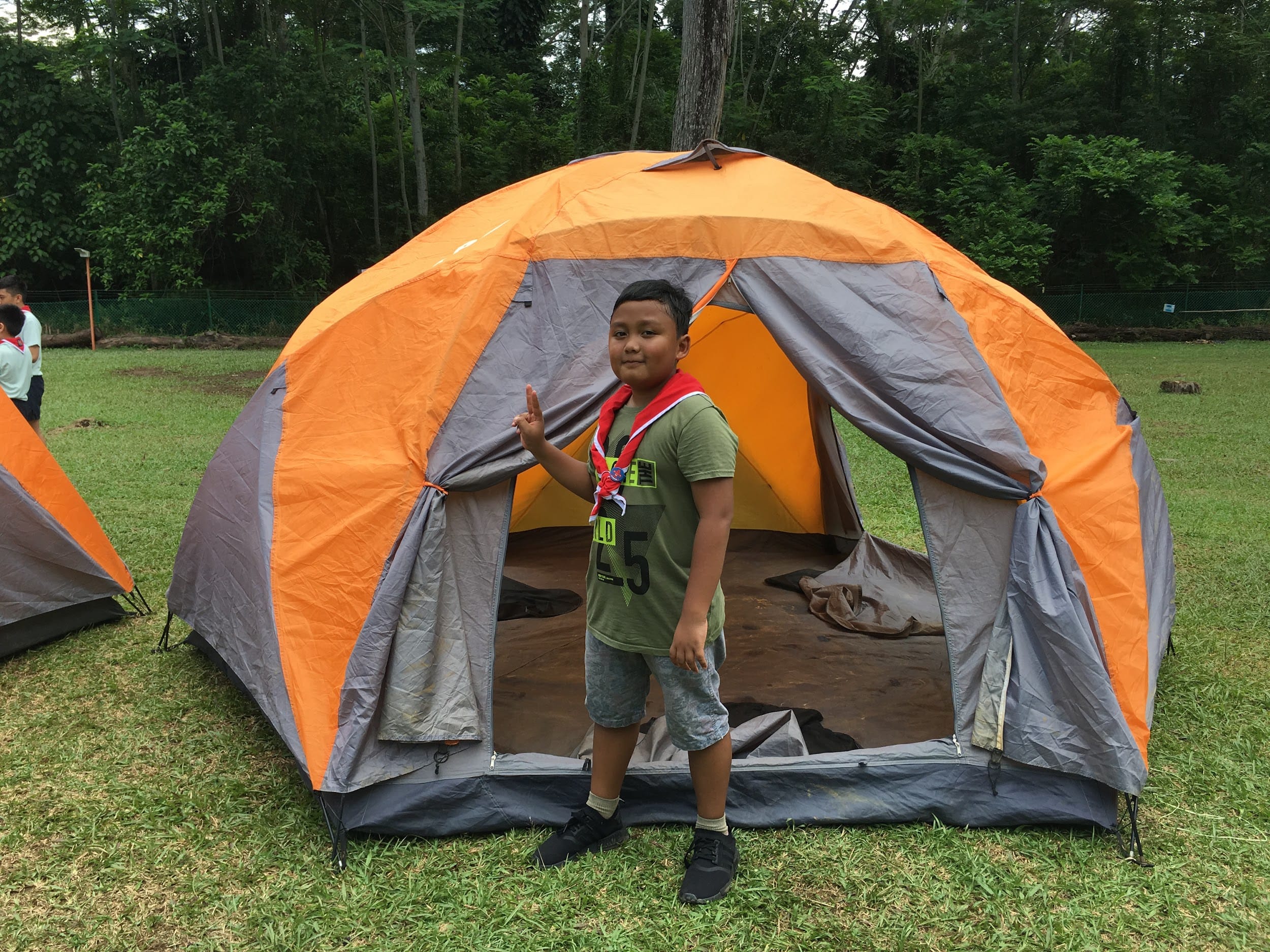 A boy in a green shirt and red neckerchief stands in front of an orange dome tent on a grassy field.
