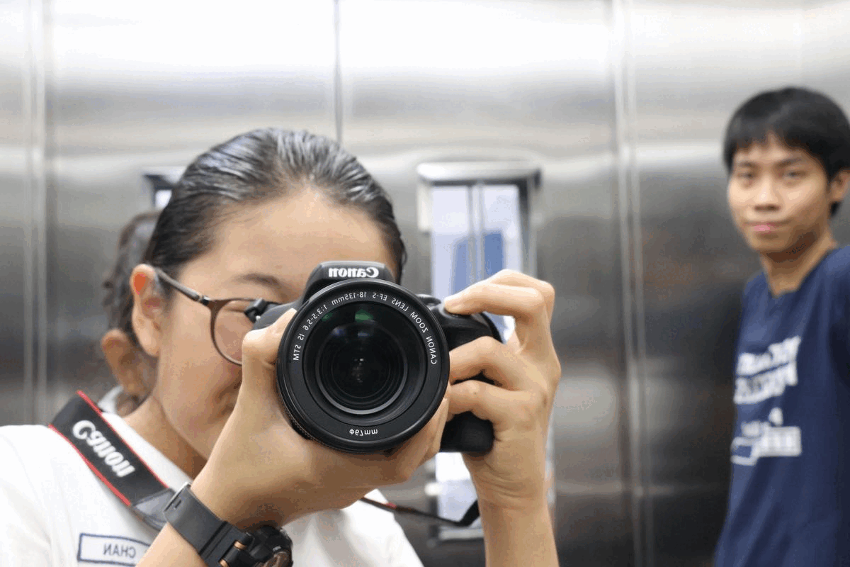 A young woman holds a Canon DSLR camera up to her eye in a mirrored elevator, with a young man visible behind her.