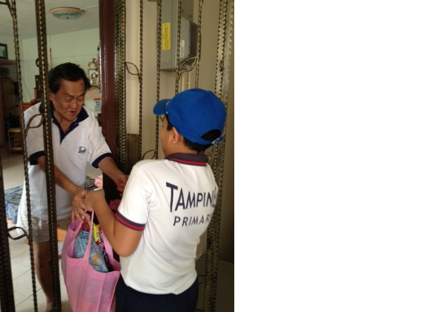 A boy in a Tampines Primary shirt and blue cap hands a bag to an older man at a gate door.