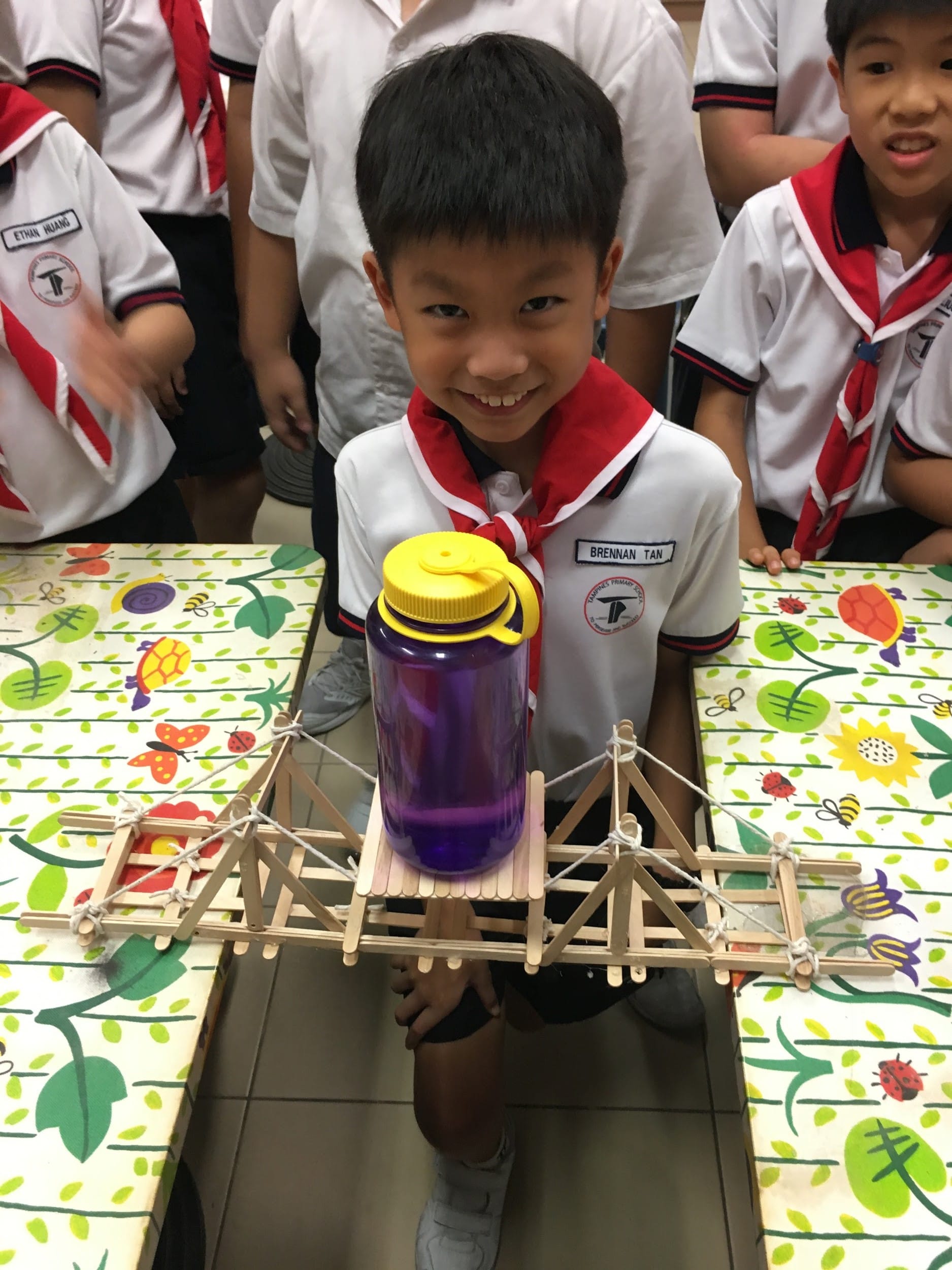 A smiling boy in school uniform holds a popsicle stick bridge supporting a purple water bottle.