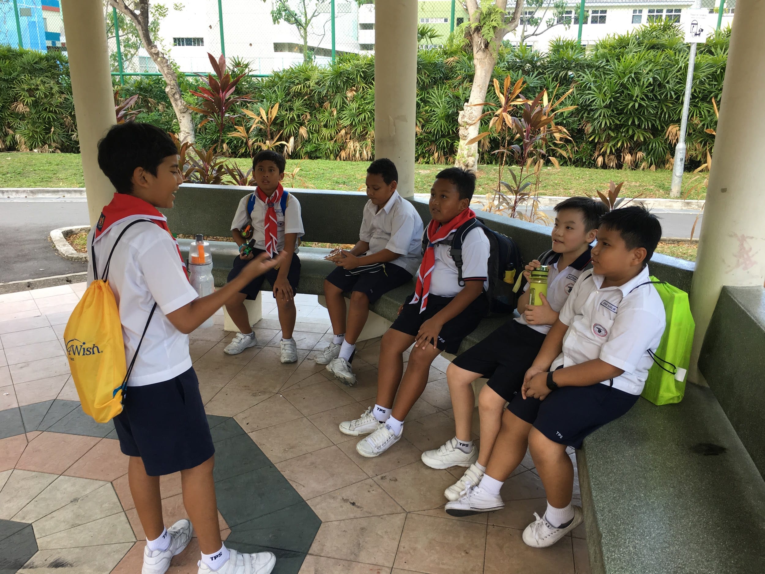 Six young boys in white school uniforms with red scarves sit and stand under a covered outdoor area, talking.