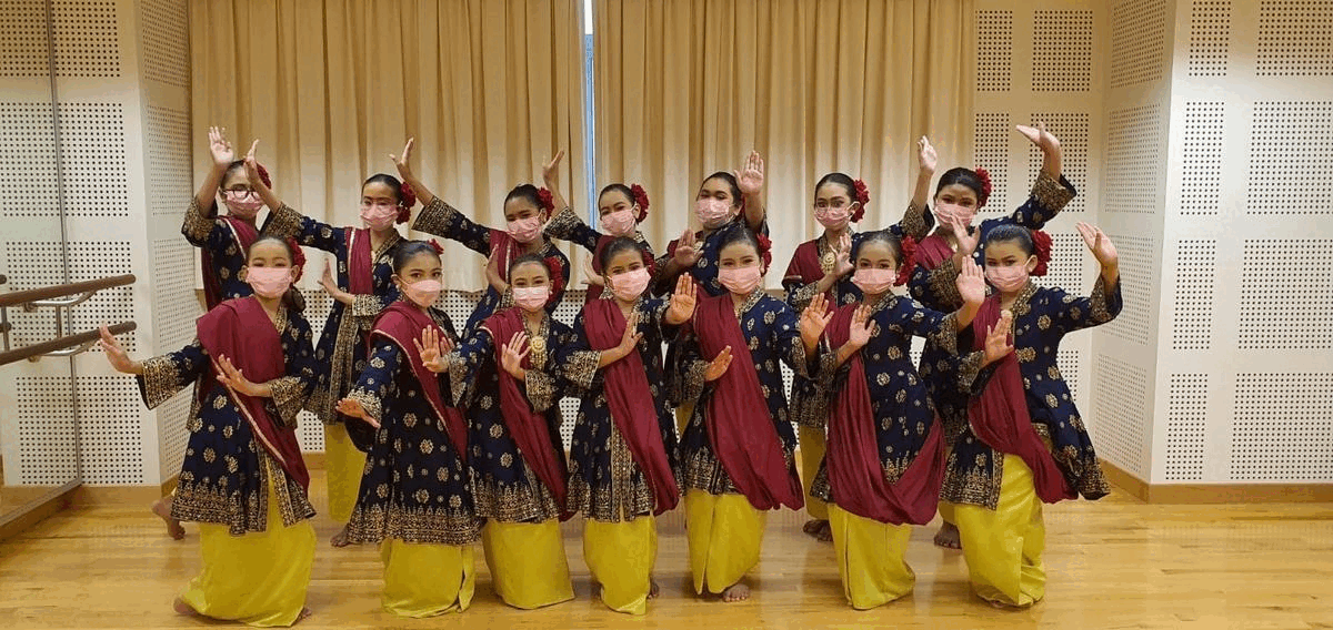 A group of young girls in blue, red, and gold traditional dance costumes and pink masks pose with raised hands.