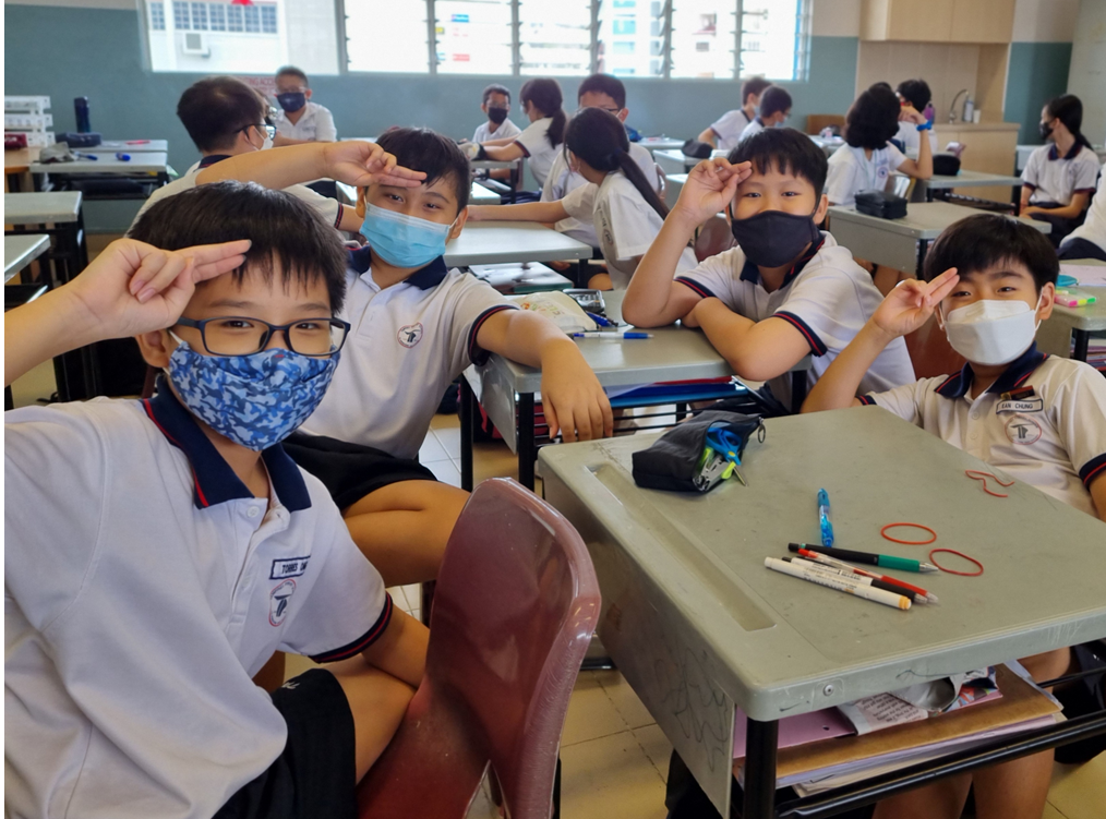 Young students in school uniforms and face masks sit at desks in a classroom, posing with peace signs.