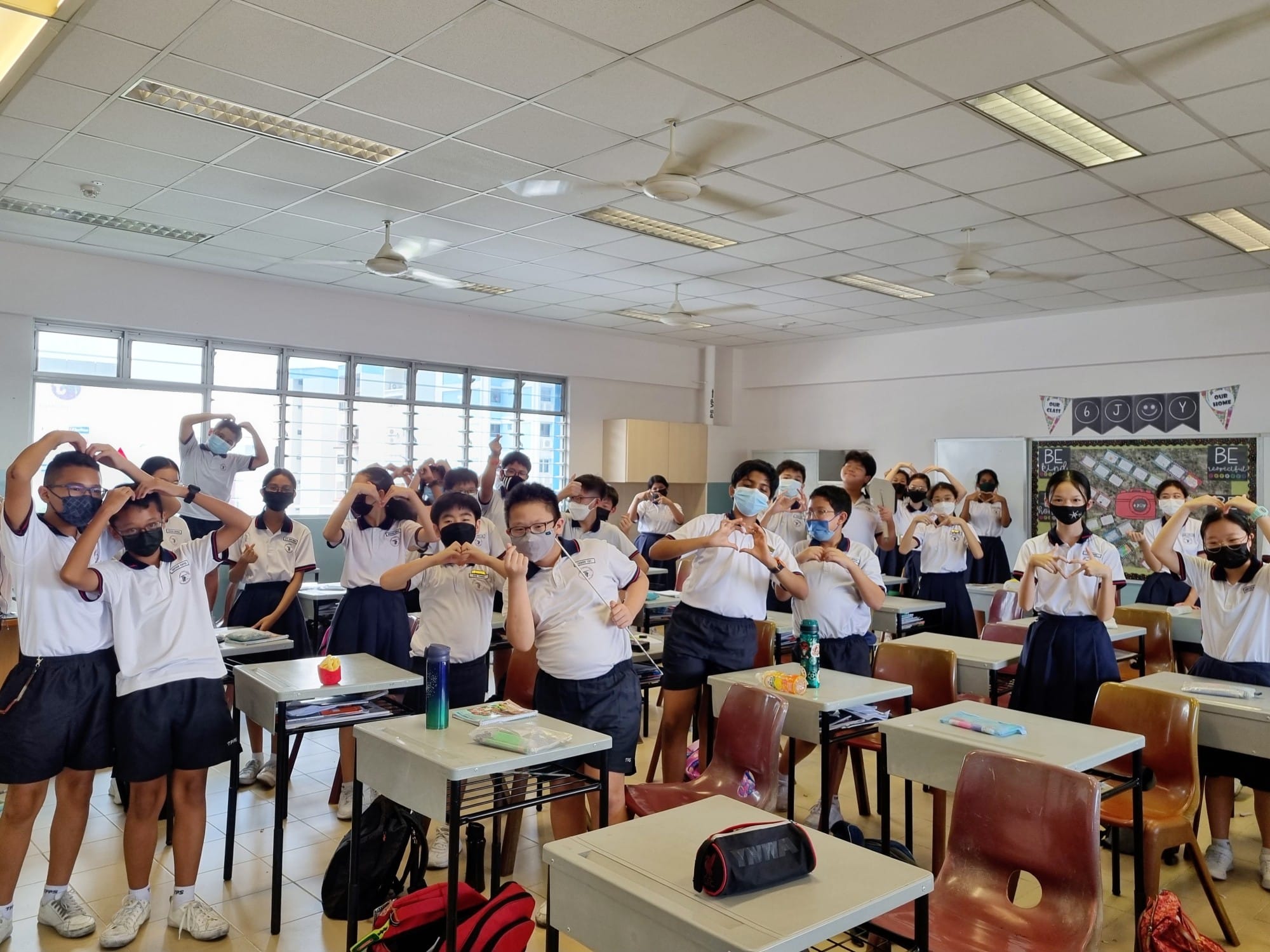 Students in uniforms and face masks stand at classroom desks making heart shapes with their hands.