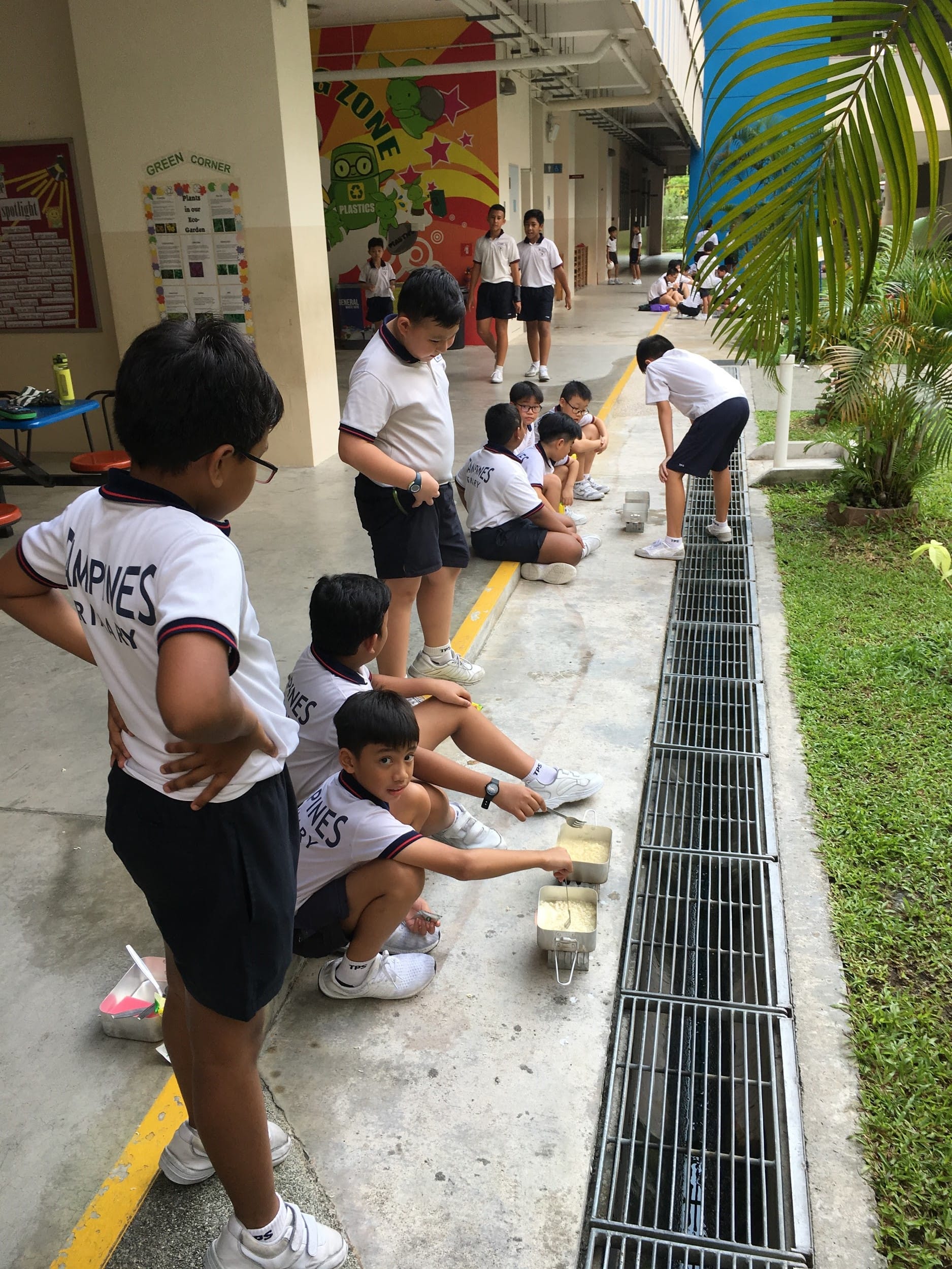 School children in white uniforms sit and stand along a drain grate in an outdoor corridor, holding small containers.