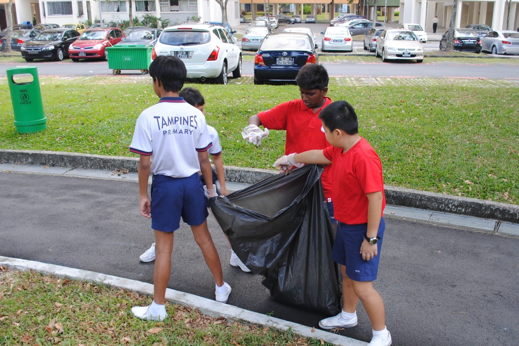 Four Tampines Primary School students in uniforms collect trash in a large black bag outdoors near a carpark.