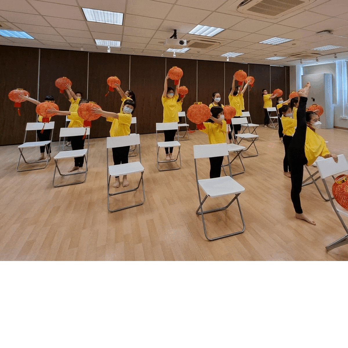 Group in yellow shirts practicing with red lanterns among rows of white chairs in a studio.