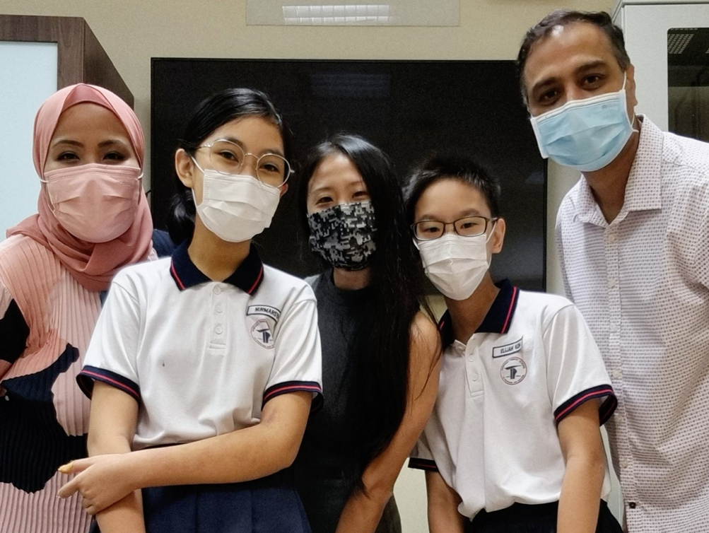Five people wearing face masks pose together, including two students in white polo uniforms and three adults.