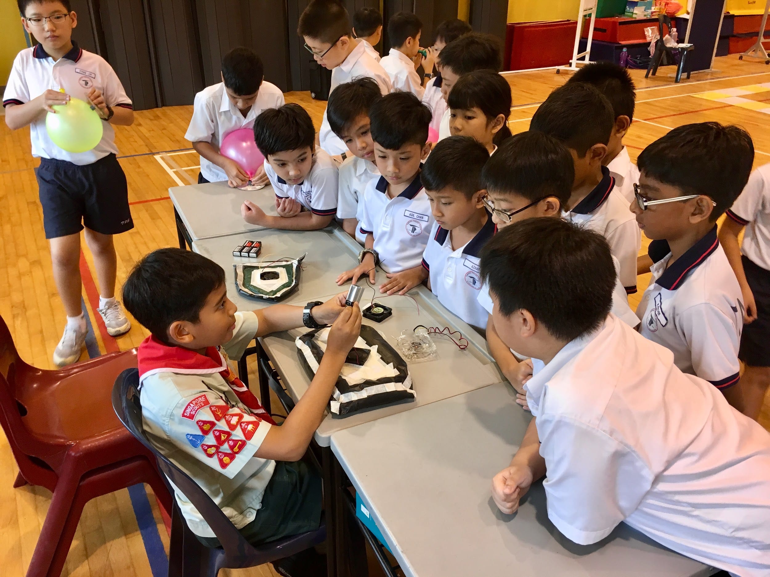 A scout boy demonstrates an electrical circuit to a group of school boys gathered around a table in a hall.