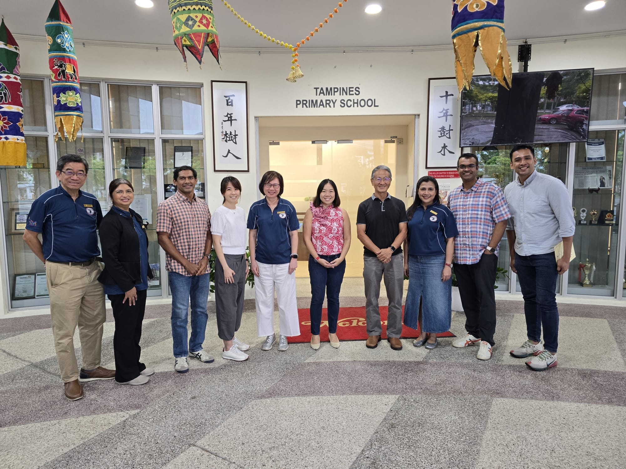 Ten adults posing together at the entrance of Tampines Primary School, decorated with colorful hanging lanterns.