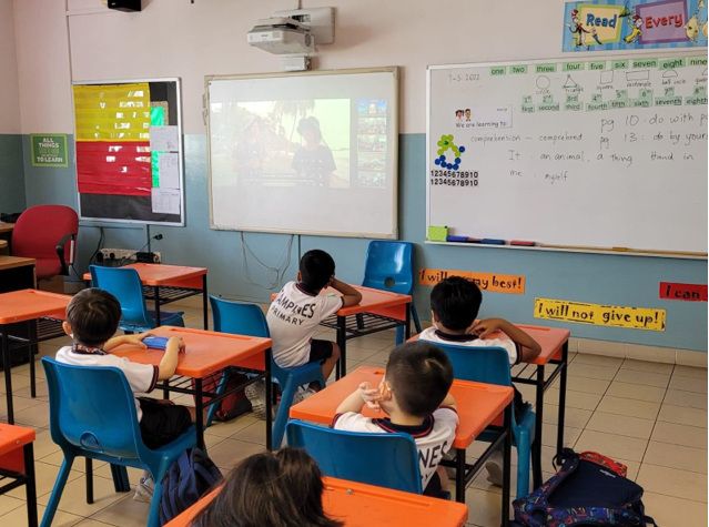 Four young students sit at orange desks watching a projected video on a whiteboard in a decorated classroom.