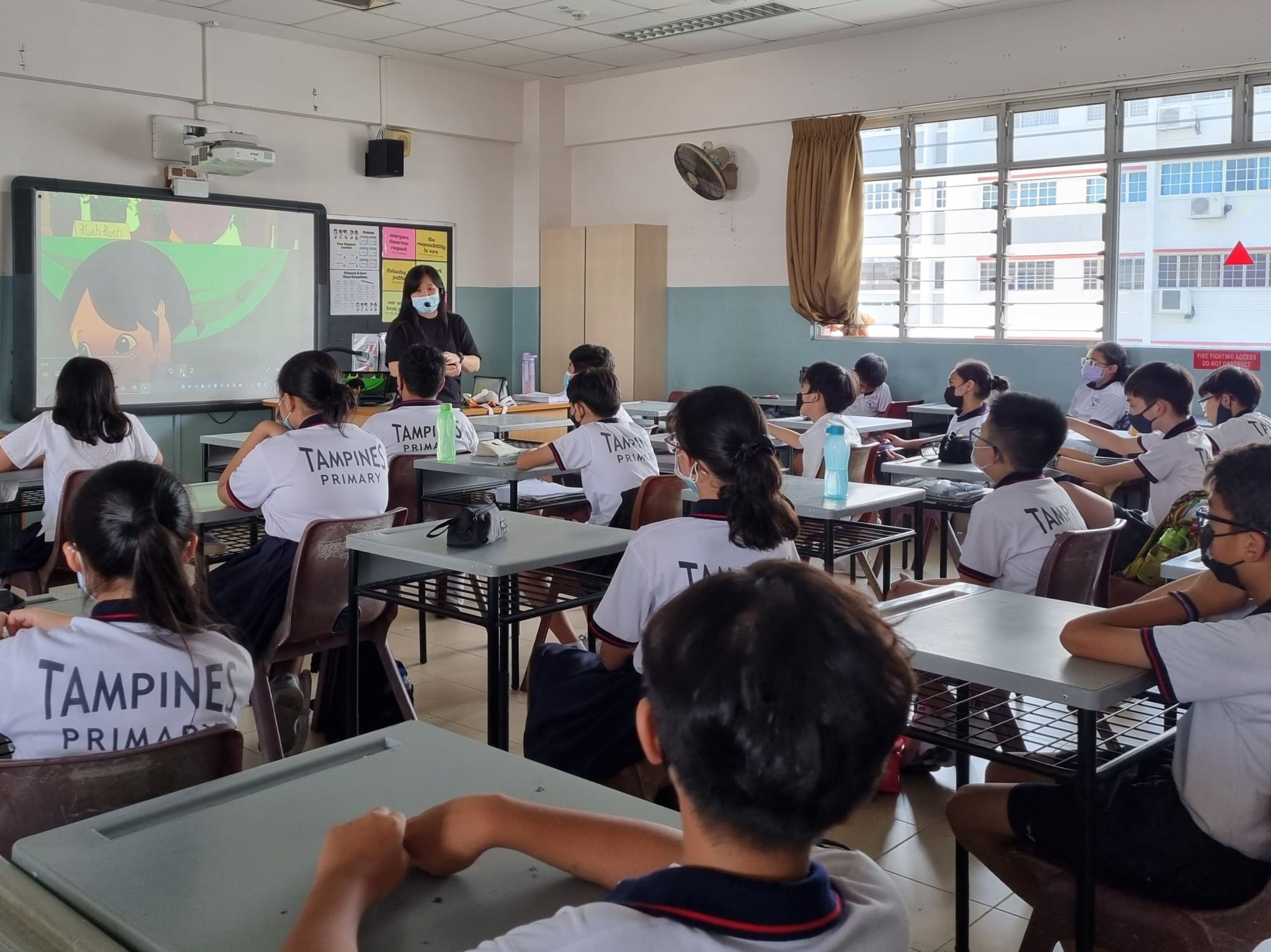 A teacher stands at the front of a Tampines Primary classroom while masked students sit at desks watching a projected screen.