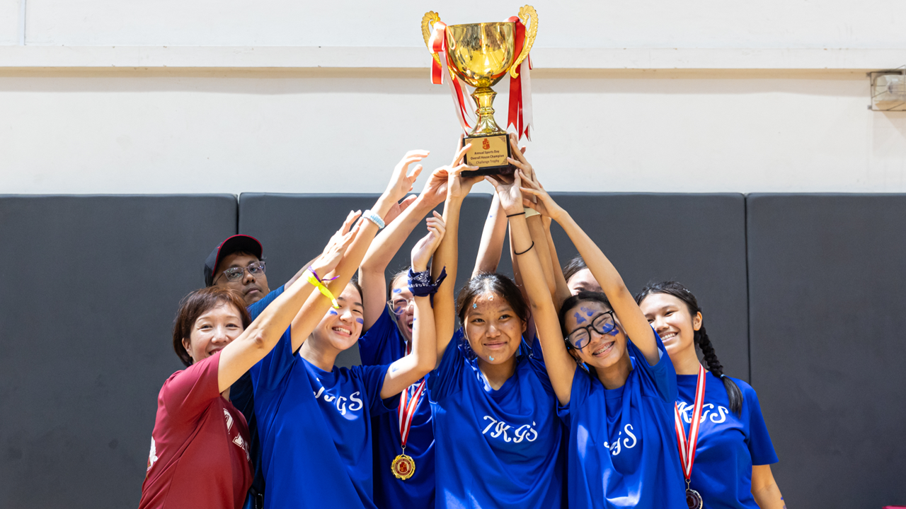 A group of students in blue TKGS shirts raise a gold trophy together, some wearing medals around their necks.