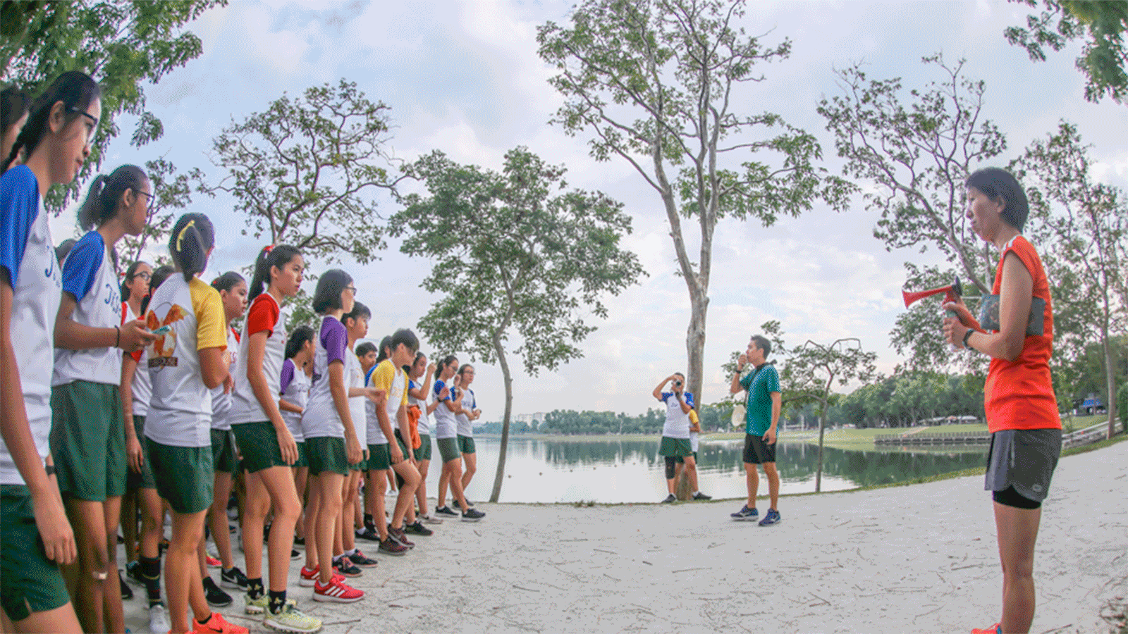 Group of students in sportswear lined up by a lake while a woman with a megaphone addresses them.